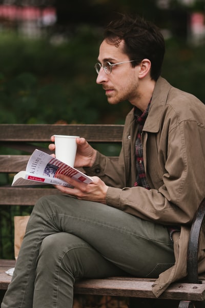 A man dressed in brown leather jacket and gray jeans sitting on the bench reading a book
